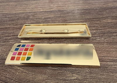 Metallskylt i blankt guld med flerfärgad emalj och ben baktill för väska